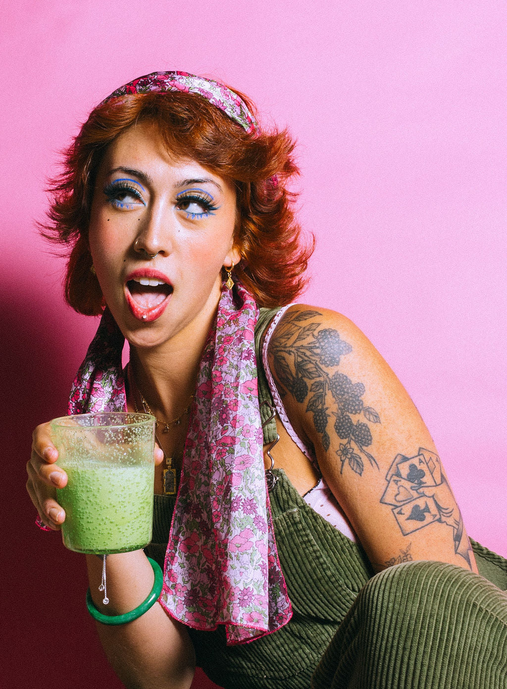 Person with tattoos and green overalls holding a matcha latte in a clear cup