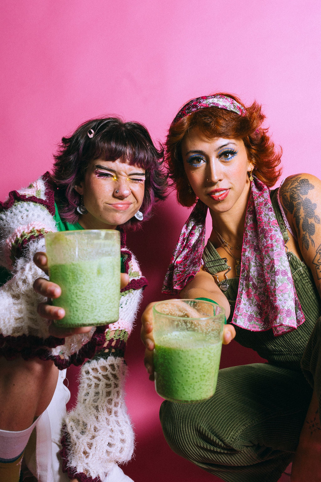 Two friends holding clear cups with matcha lattes in front of a pink background